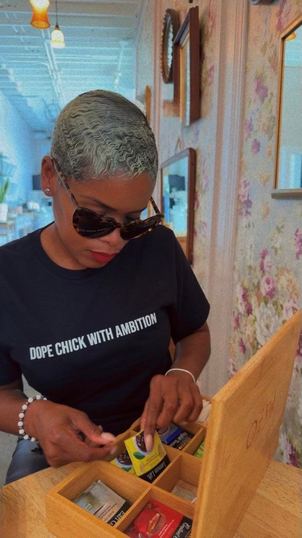 Confident Black woman in ‘Dope Chick With Ambition’ tee selects premium tea from OZTHY box in chic café, empowering lifestyle, self-care, and Black-owned fashion.
